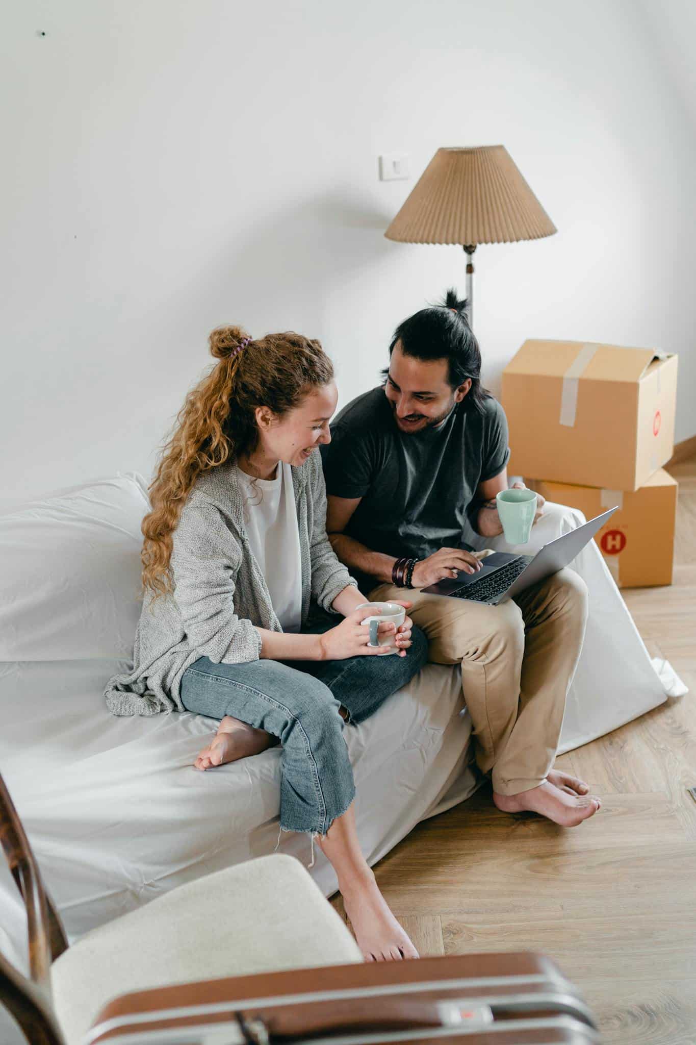 Joyful couple using a laptop and coffee in their new apartment, surrounded by moving boxes.